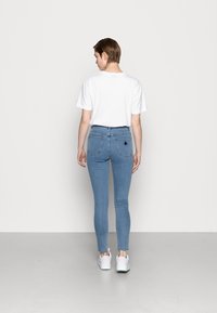 Jeans ligeiramente ajustados de cor azul claro, com uma cintura alta, apresentando dois bolsos traseiros e um detalhe em forma de triângulo. Combinados com uma t-shirt branca simples e sapatos brancos.