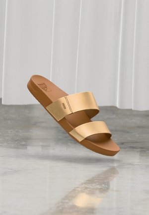 Sandalo tipo infradito color cognac con due ampie fasce in metallo dorato e suola piatta, sospeso sopra un pavimento grigio riflettente con sfondo di tende bianche.