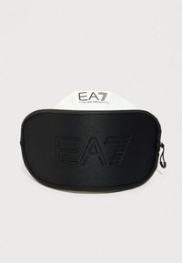 Μαύρο ορθογώνιο τσαντάκι με φερμουάρ και ανάγλυφο λογότυπο EA7 στο μπροστινό μέρος, καθώς και λευκή ετικέτα με την επωνυμία EA7 Emporio Armani πίσω από αυτό.