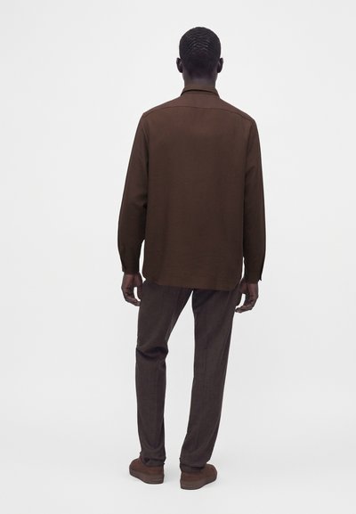 Homme debout, tourné de dos, portant une chemise marron à manches longues, un pantalon marron foncé et des chaussures marron sur un fond blanc uni.