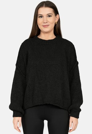 Junge Frau mit langen braunen Haaren, die einen lockeren schwarzen Strickpullover und schwarze Hosen trägt, stehend vor einem schlichten hellen Hintergrund.