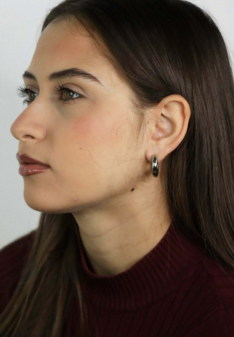 Joven con cabello largo y oscuro, vestida con un suéter de cuello alto rojo oscuro acanalado y pequeños pendientes de aro plateados, mirando hacia la izquierda en un entorno neutro.