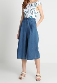 Denimové kalhoty culottes s vysokým pasem, volným střihem a širokými nohavicemi. V kombinaci s bílým topem se zelenými vzory listů. Sandály zahrnuty.