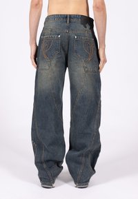 Luźne jeansy z denimu o ciemnym niebieskim odcieniu, z kontrastującymi pomarańczowymi przeszyciami na tylnej kieszeni i metalowymi detalami przy nogawkach.