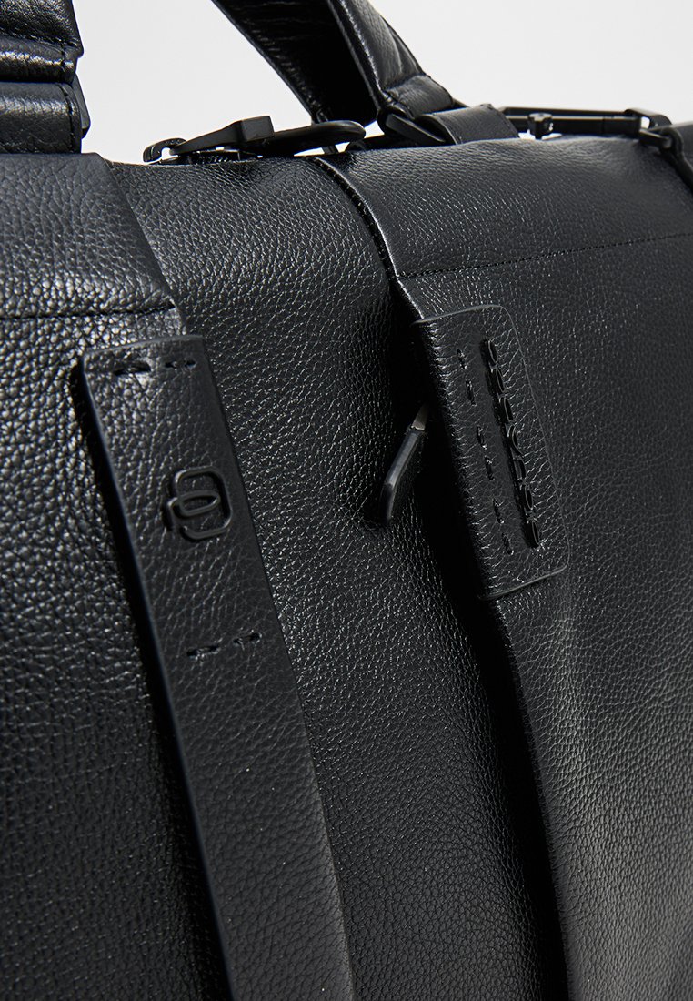 Czarna skórzana torba o teksturowanym wykończeniu, z regulowanymi paskami i minimalistycznym designem. Widoczne akcenty hardware.