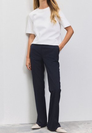 Vrouw die een effen wit T-shirt met korte mouwen, donkerblauwe wijde broek en lichtbeige loafers draagt, staand met één hand in haar zak.