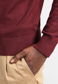 Burgundy tröja med ribbat nederkant och ärmslut, tillverkad av mjukt stickat material. I kombination med beige byxor, som visar en avslappnad passform och en avslappnad design.