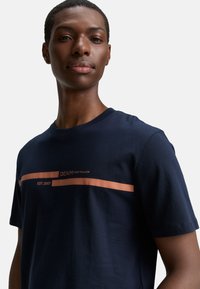 Marineblå bomulds T-shirt med korte ærmer, prydet med en vandret kobberstribe og teksten "DENIM TOM TAILOR EST. 2007" i midten.