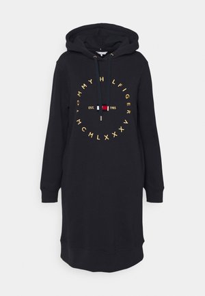 Czarna sukienka z kapturem z długimi rękawami, sznurkiem przy kapturze oraz złotym, okrągłym napisem "Tommy Hilfiger" otaczającym małe logo z flagą w kolorach czerwonym, białym i niebieskim.
