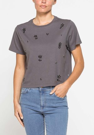 Donna che indossa una maglietta grigia a maniche corte con piccoli disegni floreali neri abbinata a jeans azzurri, una mano in tasca, su sfondo bianco.