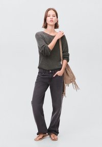 Frau trägt einen dunkelgrauen Pullover und schwarze weit geschnittene Jeans mit silbernen Nieten, hält eine beigefarbene Fransenhandtasche und steht mit einer Hand in der Tasche.