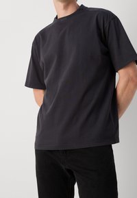 Homme portant un t-shirt noir uni à manches courtes avec un petit logo "BOSS" près du col et un pantalon noir sur un fond uni.