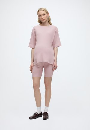 Femeie stând într-un top cu mâneci scurte din material tip rib în roz pal și pantaloni scurți asortați până la genunchi, purtând șosete albe și pantofi loafer maro închis.