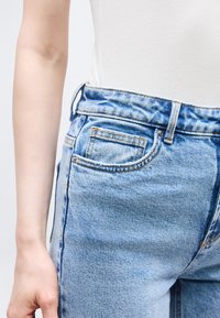 Κοντινό πλάνο ατόμου που φοράει ανοιχτό μπλε τζιν τζιν και λευκό ριμπ αμάνικο τοπ, δείχνοντας το αριστερό του χέρι και τη μέση.