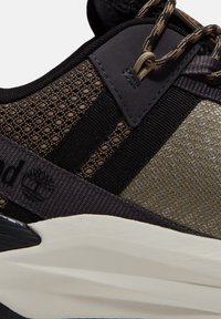 Gros plan d'une basket marron et noire montrant un tissu texturé, des lacets et une partie de la semelle blanche avec le logo visible de la marque sur le côté.
