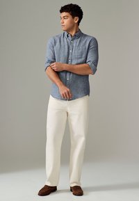 Svijetloplava košulja na kopčanje s kratkim rukavima, uparena s kremastim hlačama i smeđim cipelama. Košulja ima jedan džep na prsima.