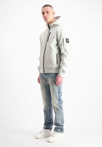 Lichtgrijze hoodie met een voorrits, twee zijzakken en een gestructureerde patch op de linkerarm, gecombineerd met vervaagde blauwe jeans en witte sneakers.