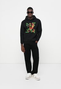 Black hoodie with a colorful graphic, paired with black pants and white sneakers. Model wearing sunglasses, standing against a white background.