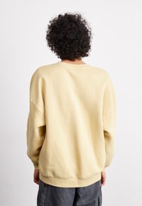 Osoba s kudrnatými tmavými vlasy, oblečená v volném béžovém svetru a šedých cargo kalhotách, stojící čelem k jednoduchému bílému pozadí.