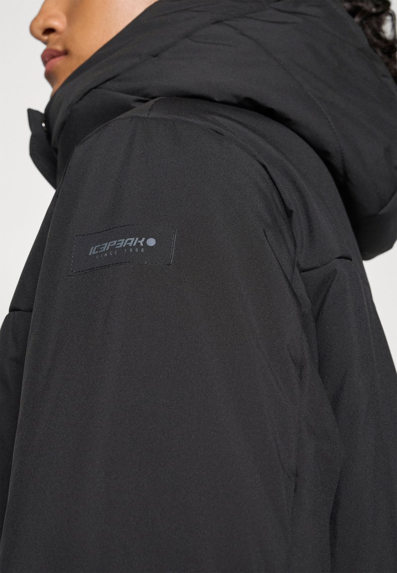 Nahaufnahme einer Person, die eine schwarze gepolsterte Jacke mit großer Kapuze und sichtbarem "Icepeak"-Logo am oberen Ärmel trägt.