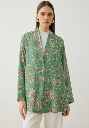 Femme portant un blazer vert avec des motifs floraux marron et blancs sur un top blanc et un pantalon blanc, sur un fond clair uni.