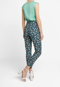 Green sleeveless top paired with patterned tapered trousers featuring a blue, pink, and green floral design, complemented by white block-heel sandals.