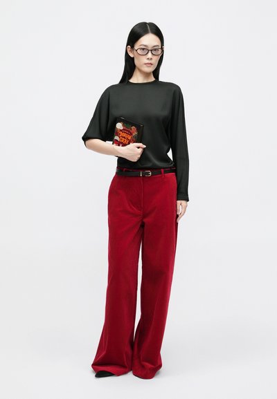 Top nero a maniche corte, pantaloni di velluto a coste rossi larghi con una cintura, in mano un libro. Occhiali chiari, capelli scuri e lunghi, accessori minimi.