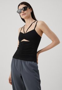 Top negro sin mangas con frente fruncido, detalle de corte y tirantes delgados. Combinado con pantalones grises sueltos y brillantes.