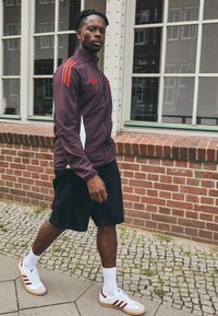 Giacca sportiva color marrone con strisce rosse, dettagli bianchi e chiusura con zip, abbinata a pantaloni corti neri e sneakers bianche con strisce rosse.