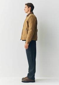 Brown, lightweight jacket with a zip closure and side pockets, paired with dark pants and brown boots with orange laces. Side profile view.