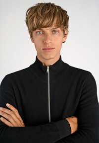 Svart zip-up-tröja med strukturerad tyg, hög krage och ribbade ärmslut. Modellen poserar med armarna i kors och visar plaggpassformen.