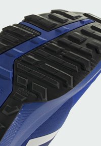 Baskets de sport bleues avec une semelle en caoutchouc texturée à profondeurs de crans, des accents mats et brillants, et un capuchon renforcé.