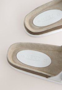 Sandalias beige de ante con plantillas contorneadas, parches ovalados blancos con relieve "NEXT FOREVER COMFORT" y suelas exteriores blancas sobre un fondo neutro.