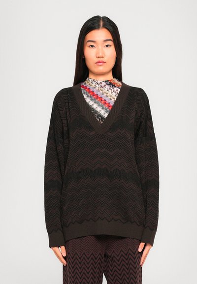 Jonge vrouw met lang, steil haar draagt een donkere V-halstriktrui met zigzagpatroon, bijpassende broek en een meerkleurig topje eronder.