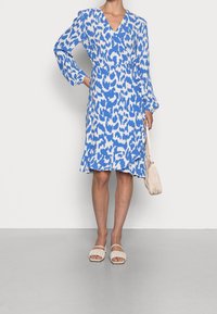 Vestido de leopardo azul y blanco con escote en V, mangas largas y dobladillo con volantes; combinado con sandalias de tela color crema y un bolso texturizado.