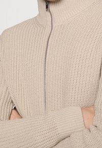 Beige pletený svetr s vysokým límcem, středovým zipem, žebrovanou strukturou a přiléhavými manžetami, zdůrazňující těsný a strukturovaný design.