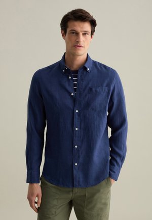Navy blue button-up shirt made of linen, featuring a single chest pocket and a casual collar, layered over a striped shirt.