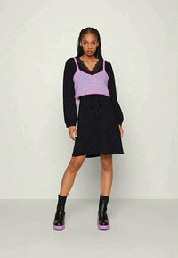 Vestido preto com cintura ajustável, sobreposto a uma blusa sem mangas em xadrez fúcsia com acabamento em renda. Botas de plataforma pretas com solas roxas.