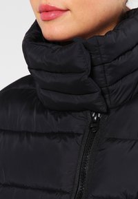Primer plano de labios y barbilla con una chaqueta tipo abrigo acolchado negra con un alto cuello acolchado y un cierre oscuro visible.