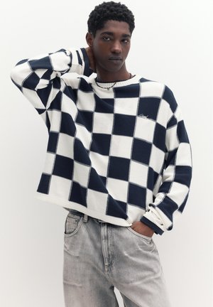 Mladý muž s tmavými kučeravými vlasmi, oblečený v čierno-bielom károvanom svetri a svetlošedých džínsoch, pózujúci s jednou rukou za krkom.