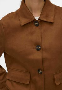 Veste en daim marron avec un col à revers, des boutons sur le devant et deux poches plaquées. Le tissu a un aspect doux et texturé.