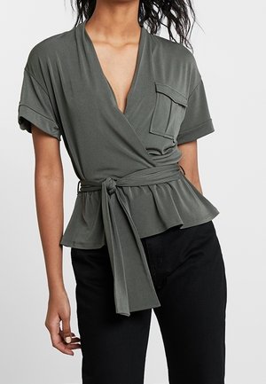 Frau trägt eine olivgrüne, kurzärmelige Wickelbluse mit Gürtel und Brusttasche, kombiniert mit schwarzen Hosen.
