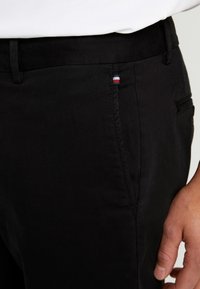 Detailní pohled na kapsu černých kalhot s malou přišitou etiketou s červenými, bílými a modrými pruhy na osobě oblečené v bílém topu.