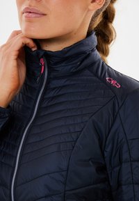 Mørkeblå isoleret jakke med høj krave, quiltet design, grå lynlås og rosa brandingdetalje på skulderen. Glat tekstur.