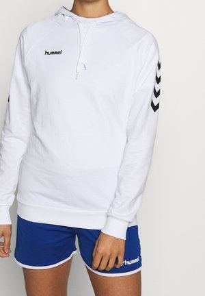 Osoba ubrana w biały kaptur hummel z czarnymi wzorami typu chevron na rękawach oraz niebieskie sportowe szorty hummel na jednolitym tle.