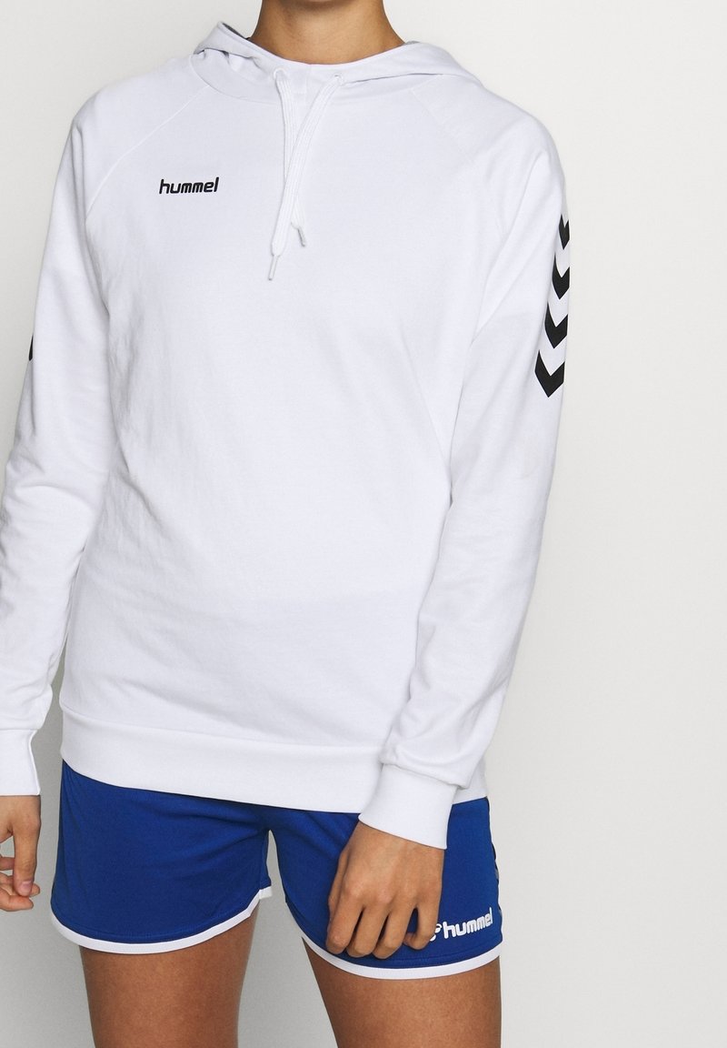 Personne portant un sweat à capuche blanc hummel avec des chevrons noirs sur les manches et un short de sport bleu hummel sur un fond uni.