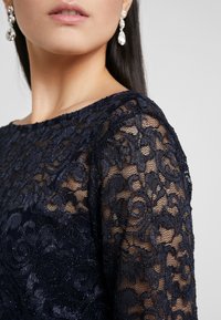 Námorně modré krajkové šaty s květinovým vzorem, třpytivou texturou a lodním výstřihem. Doplnené visacími náušnicemi.
