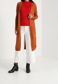 Dlouhý otevřený cardigan v oranžové ribované textuře, vrstvený přes červený rolák a bílé kalhoty s širokými nohavicemi. Černé kotníkové boty dotvářejí celý outfit.