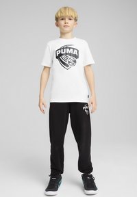 Camiseta blanca de algodón con el logo negro de "PUMA", combinada con pantalones de chándal negros que presentan un pequeño logo. Zapatillas deportivas negras con acentos en azul verdoso.