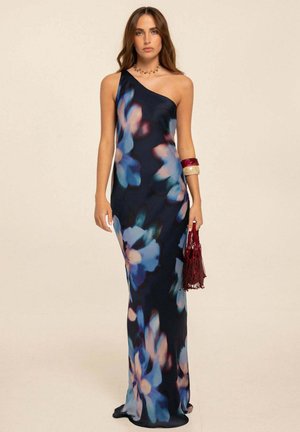 Mujer que lleva un vestido maxi de un hombro oscuro con un gran estampado floral azul, sosteniendo un bolso rojo con borlas y llevando pulseras gruesas.